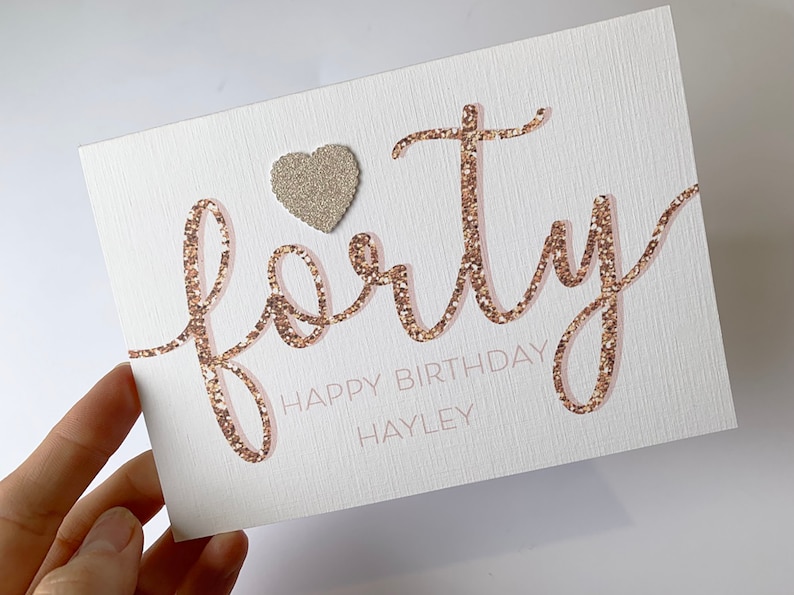 May include: A white card with the word "forty" written in gold glitter. The word "happy birthday" is written below the word "forty" in black text. The name "Hayley" is written below the word "happy birthday" in black text. A small gold glitter heart is in the upper left corner of the card.