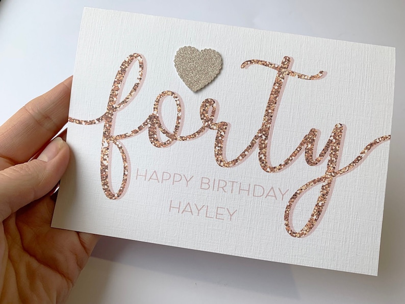 May include: A white card with the word "forty" written in pink and gold glitter. The word "happy birthday" is written below in pink and the name "Hayley" is written below that in pink. A small silver glitter heart is above the word "forty".
