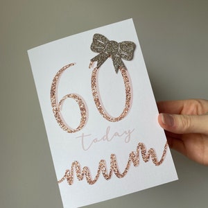 May include: A white card with the number 60 in rose gold glitter, the word "today" in pink script, and the word "mum" in a rose gold glitter script. A silver glitter bow is adhered to the top of the card.