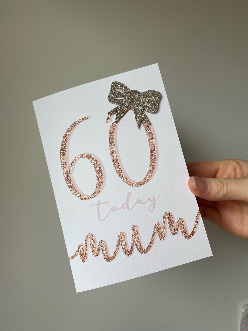May include: A white card with the number 60 in rose gold glitter, the word "today" in pink script, and the word "mum" in a rose gold glitter script. A silver glitter bow is adhered to the top of the card.
