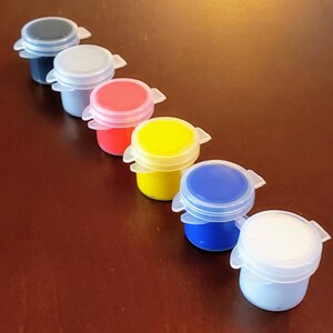 May include: Seven small paint pots with lids, lined up in a row on a wooden surface. The pots are filled with paint in the colors gray, light blue, red, yellow, blue, white, and black.