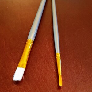 May include: Two paint brushes with silver handles and gold ferrules. The brush on the left has a flat, white bristle head. The brush on the right has a round, white bristle head.