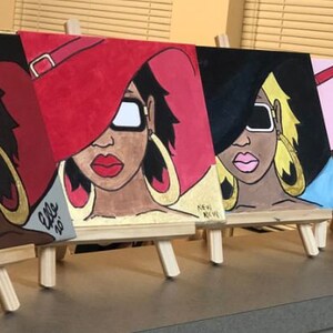 May include: Four small canvases with paintings of women wearing hats and sunglasses. The paintings are on easels and are displayed on a table. The women are all wearing different colored hats and sunglasses. The paintings are all in a similar style and are very colorful.