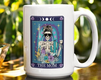 May include: White ceramic coffee mug with a colorful illustration of a skeleton wearing sunglasses and a bandana, holding a glass of wine. The illustration is surrounded by cacti and flowers. The text "THE MOM" is printed below the illustration.