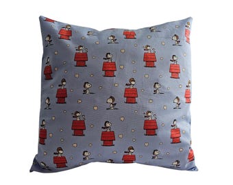 Puede incluir: Una almohada azul con un patrón repetido de personajes de Snoopy volando aviones y casas de perro rojas.