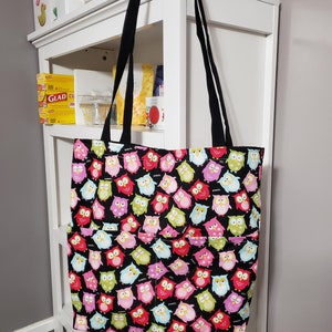 May include: A black tote bag with a colorful owl print. The bag has two black handles and two pockets on the front.