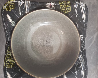 May include: A gray ceramic bowl sits on a black and white Star Wars themed fabric hot pad. The pad features the text "Star Wars" and images of X-Wing starfighters.