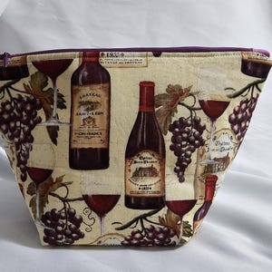 Puede incluir: Un neceser de tela beige con temática de vino. La bolsa presenta imágenes de botellas de vino, copas de vino tinto y racimos de uvas. El borde superior está adornado con tela morada y tiene una cremallera morada.