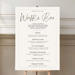 Könnte beinhalten: Ein cremefarbenes Schild auf einer Staffelei mit der Aufschrift "Make Your Own Waffle Bar". Das Schild listet Waffel-Toppings und Dessert-Kombinationen wie S'mores, Berries & Cream und Ice Cream Sundae auf. Der Text ist in einer eleganten Schriftart.
