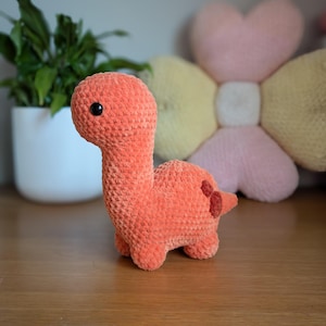 Pu&ograve; includere: Un giocattolo di dinosauro di crochet arancione con occhi neri. Il dinosauro &egrave; seduto su una superficie di legno.