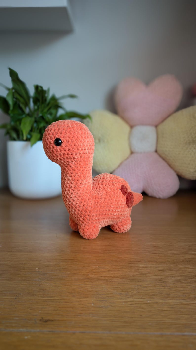 Pu&ograve; includere: Un giocattolo di dinosauro di crochet arancione con occhi neri. Il dinosauro &egrave; seduto su una superficie di legno.
