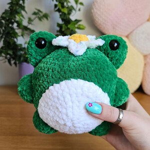 May include: A handmade green plush frog with a white belly and a daisy on its head. The frog has large black eyes and is made of soft, textured yarn. A charming, whimsical stuffed animal.