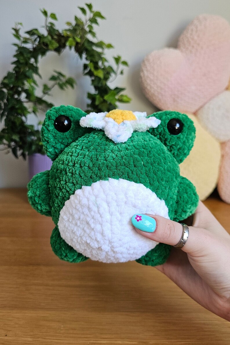 May include: A handmade green plush frog with a white belly and a daisy on its head. The frog has large black eyes and is made of soft, textured yarn. A charming, whimsical stuffed animal.