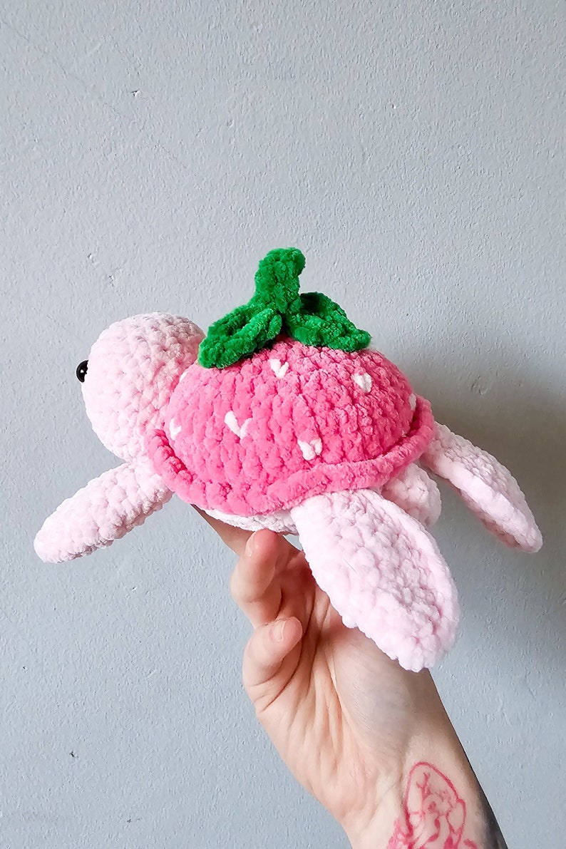 May include: A pink crocheted turtle with a strawberry-shaped shell and green leaves on top.