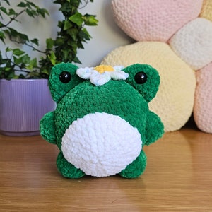May include: A handmade green and white crochet frog plushie with a daisy on its head. The frog has large black eyes and is sitting on a wooden surface. A purple pot with greenery is in the background.