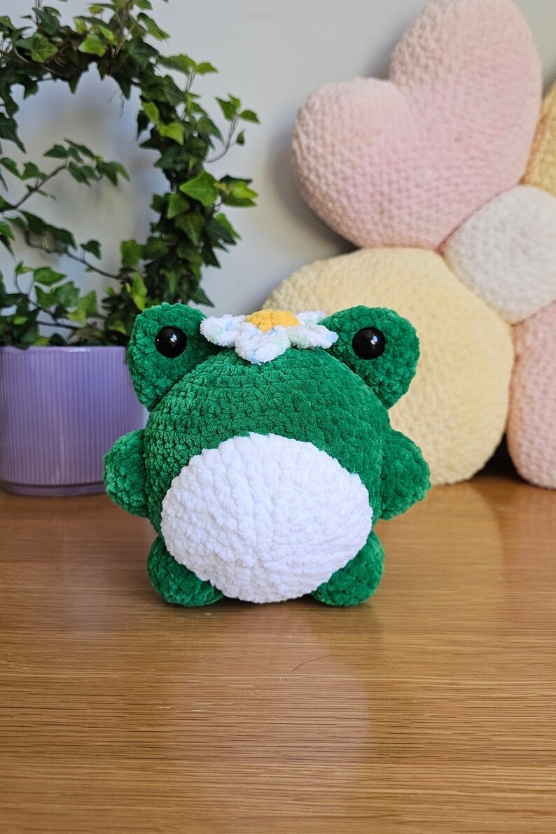 May include: A handmade green and white crochet frog plushie with a daisy on its head. The frog has large black eyes and is sitting on a wooden surface. A purple pot with greenery is in the background.