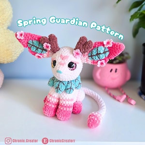 May include: A handmade crochet creature with pink and white fur, large eyes, and decorative wings. The creature has a blue and green collar and is adorned with small flowers. The text "Spring Guardian Pattern" is visible above the creature.