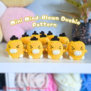 May include: A collection of small, yellow, crocheted duck figures with black hair tufts, white beaks, and embroidered eyes. The text 'Mini Mind-Blown Duckie Pattern' is displayed above the figures, with social media handles at the bottom.
