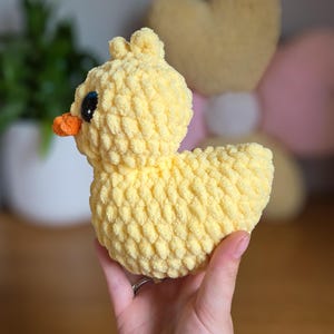 May include: A handmade, yellow crochet duck with an orange beak and black eyes. The plush duck has a textured, fluffy appearance. The duck is a soft toy, likely for children, and is held in a person's hand.