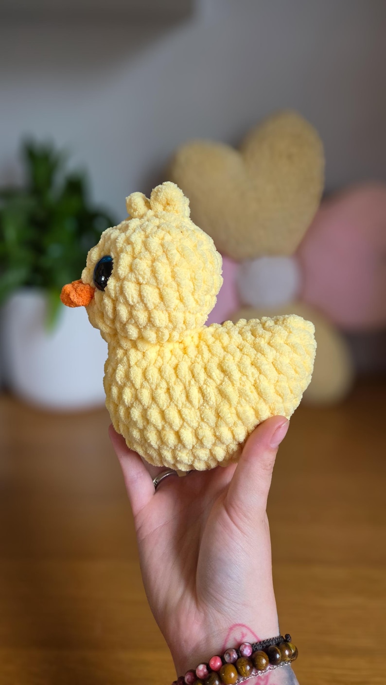May include: A handmade, yellow crochet duck with an orange beak and black eyes. The plush duck has a textured, fluffy appearance. The duck is a soft toy, likely for children, and is held in a person's hand.