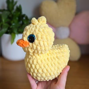 May include: A handmade yellow crochet duck with an orange beak and large blue eyes. The plush toy is soft and textured, ideal for a child's room or nursery. The duck is a charming, whimsical creation.