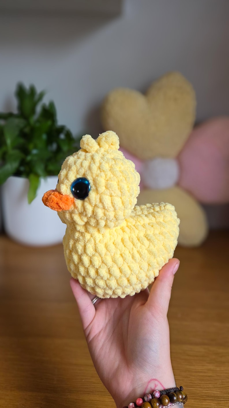 May include: A handmade yellow crochet duck with an orange beak and large blue eyes. The plush toy is soft and textured, ideal for a child's room or nursery. The duck is a charming, whimsical creation.