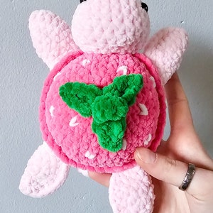 May include: A pink crocheted turtle with a strawberry design on its shell. The strawberry has white seeds and a green leaf.