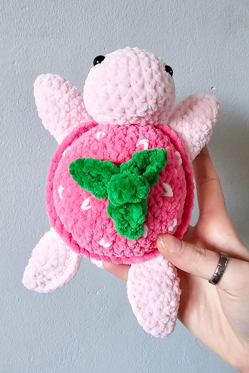 May include: A pink crocheted turtle with a strawberry design on its shell. The strawberry has white seeds and a green leaf.