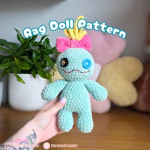 May include: A handmade, light blue rag doll with a pink bow, yellow accents, and button eyes. The doll has a stitched mouth and is held in front of a plant. The text "Rag Doll Pattern" is displayed above the doll.