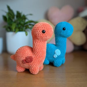 Pu&ograve; includere: Due giocattoli di dinosauro all'uncinetto, uno arancione e uno blu, con occhi neri. I giocattoli sono seduti su una superficie di legno.