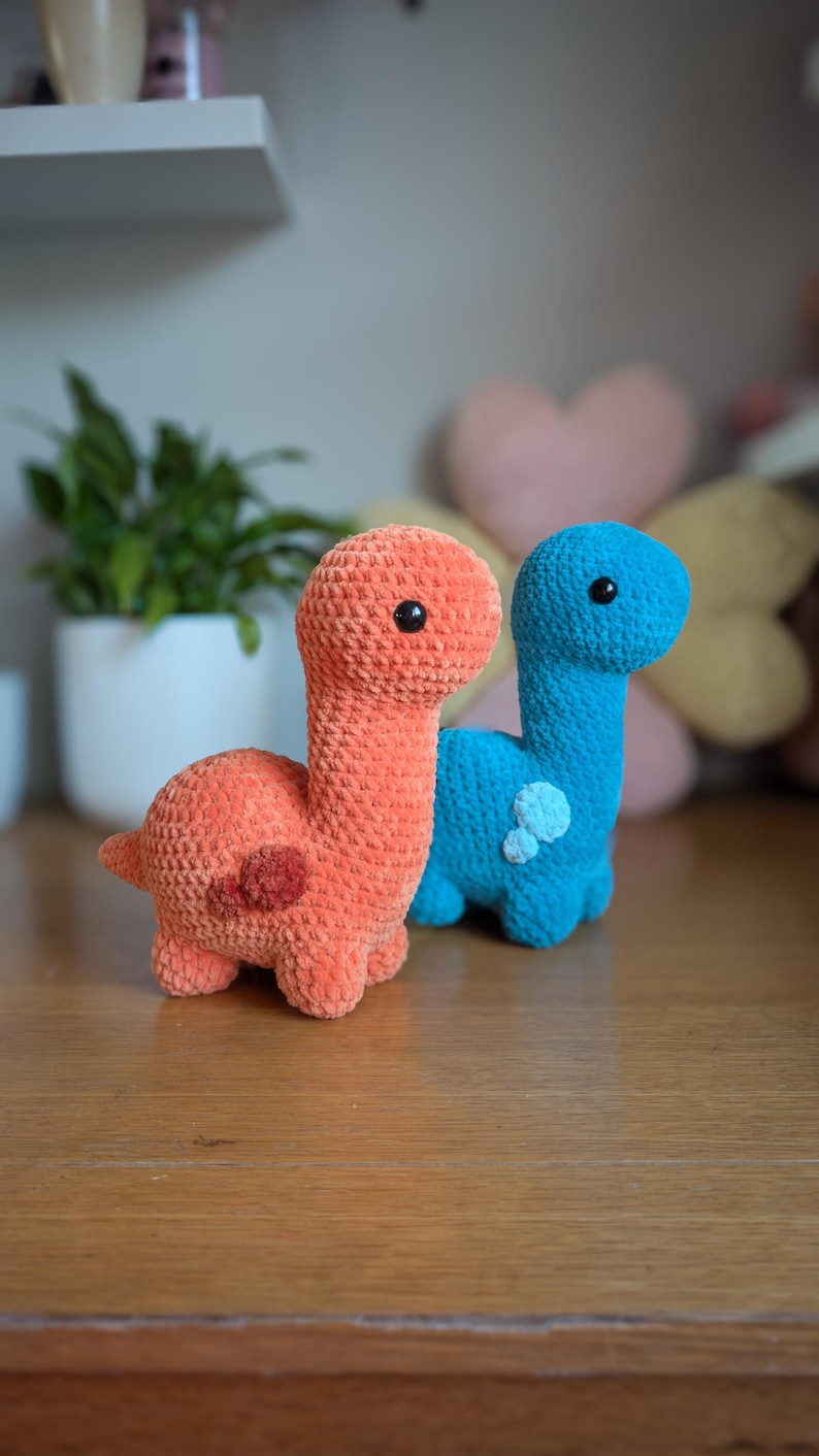 Pu&ograve; includere: Due giocattoli di dinosauro all'uncinetto, uno arancione e uno blu, con occhi neri. I giocattoli sono seduti su una superficie di legno.
