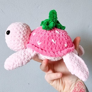 May include: A pink crocheted turtle with a strawberry shell and green leaves on top. The turtle is made of soft yarn and has a cute, friendly expression.