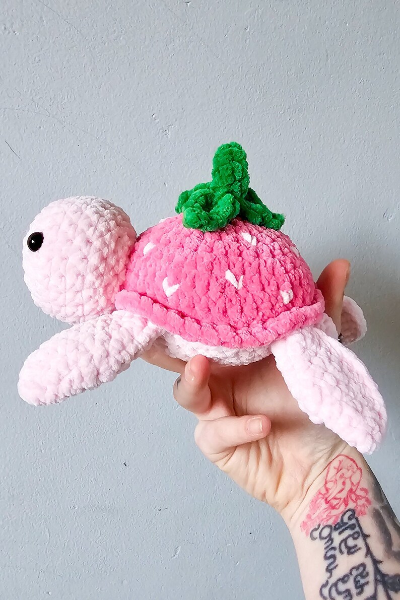 May include: A pink crocheted turtle with a strawberry shell and green leaves on top. The turtle is made of soft yarn and has a cute, friendly expression.