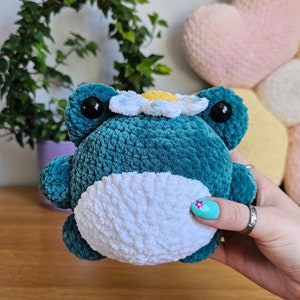 May include: A teal crocheted frog plushie with a white belly and a daisy crown. The frog has large black eyes and is held in a person's hand. The plushie is on a wooden surface.