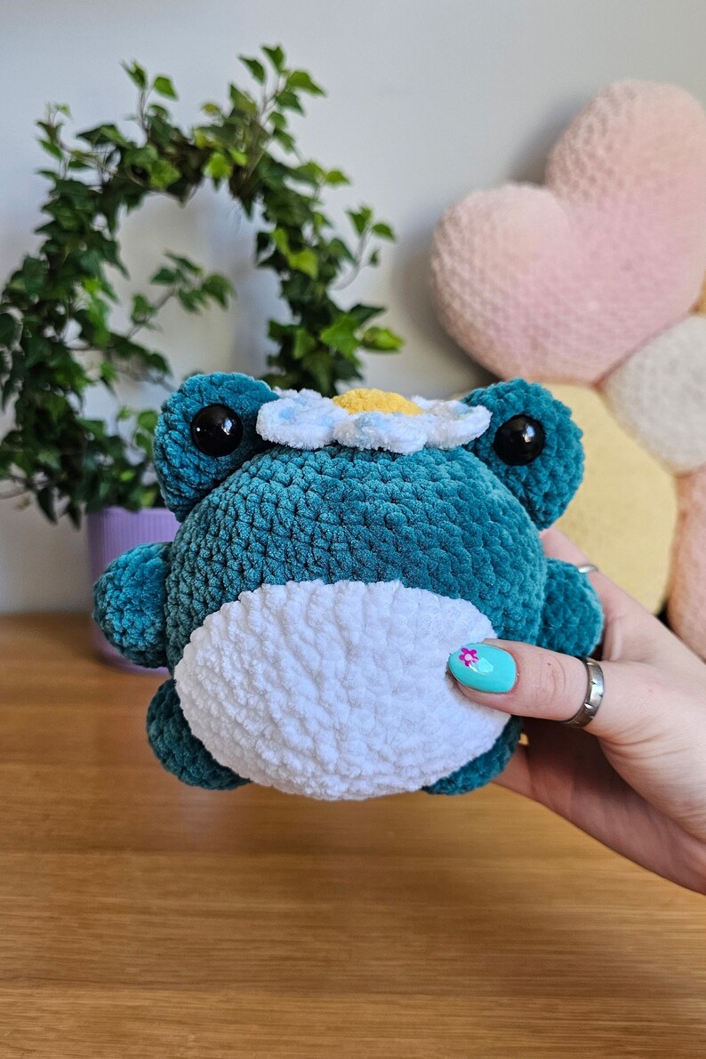 May include: A teal crocheted frog plushie with a white belly and a daisy crown. The frog has large black eyes and is held in a person's hand. The plushie is on a wooden surface.