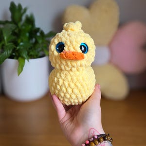 May include: A handmade yellow crochet duck with large blue eyes and an orange beak. The plush toy is soft and textured, ideal for a nursery or as a decorative item. The duck is held in a hand.