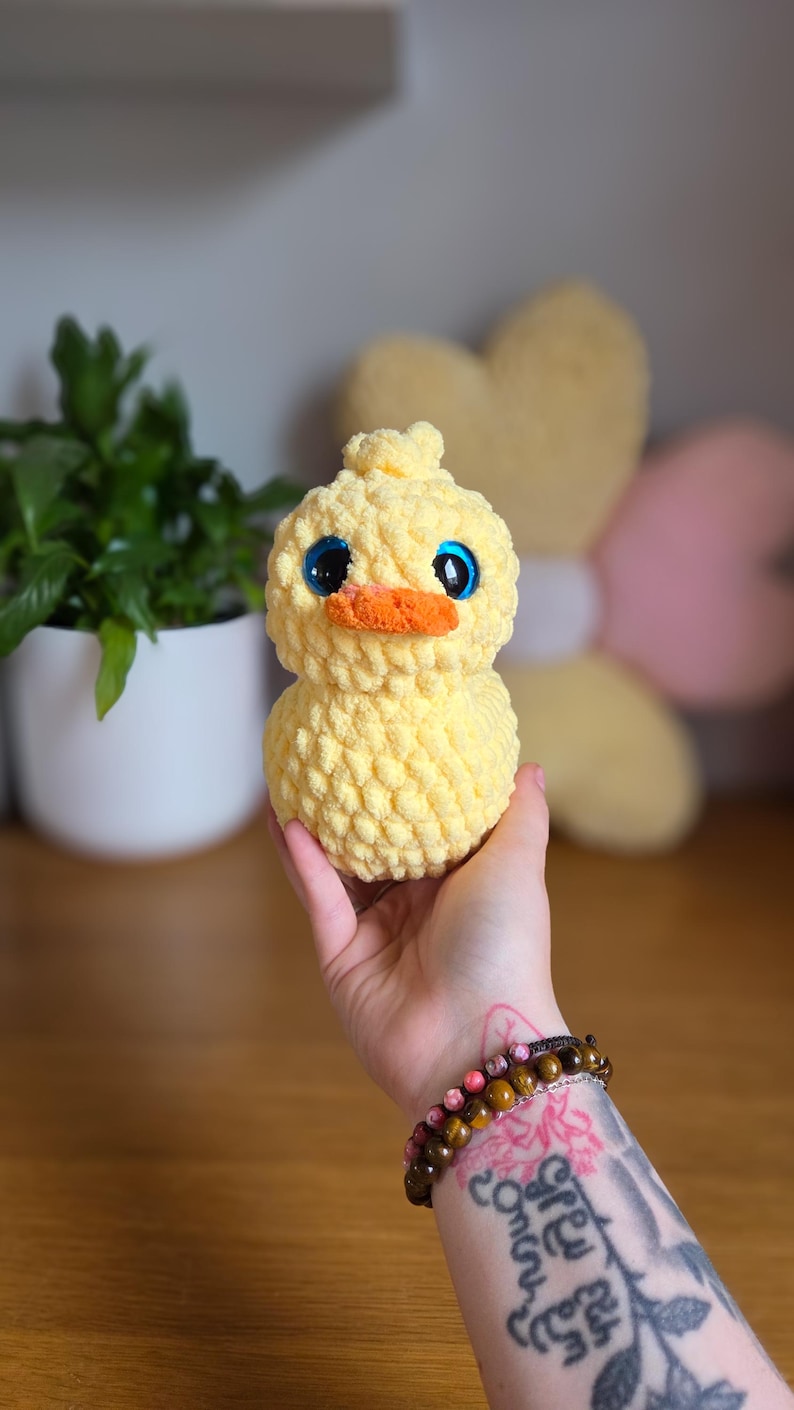 May include: A handmade yellow crochet duck with large blue eyes and an orange beak. The plush toy is soft and textured, ideal for a nursery or as a decorative item. The duck is held in a hand.