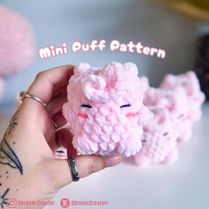 May include: A small, light pink crocheted plush toy with a textured puff stitch pattern. The toy features embroidered dark eyes and small pink cheeks. Above the plush, the text reads Mini Puff Pattern in a white, rounded font style.
