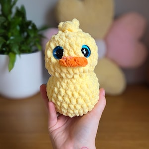 May include: A yellow crochet duckling with large blue eyes and an orange beak. The soft toy is held in a hand, highlighting its textured, rounded form. The duckling is a charming, whimsical item.