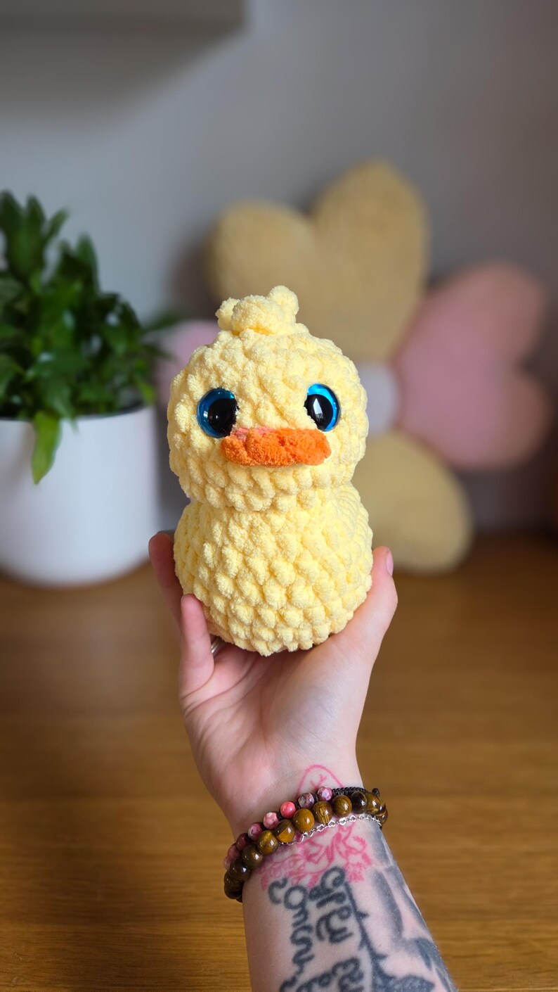 May include: A yellow crochet duckling with large blue eyes and an orange beak. The soft toy is held in a hand, highlighting its textured, rounded form. The duckling is a charming, whimsical item.
