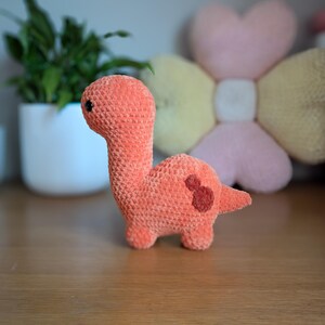 Pu&ograve; includere: Un giocattolo di dinosauro di crochet arancione con occhi neri. Il dinosauro &egrave; in piedi su una superficie di legno.