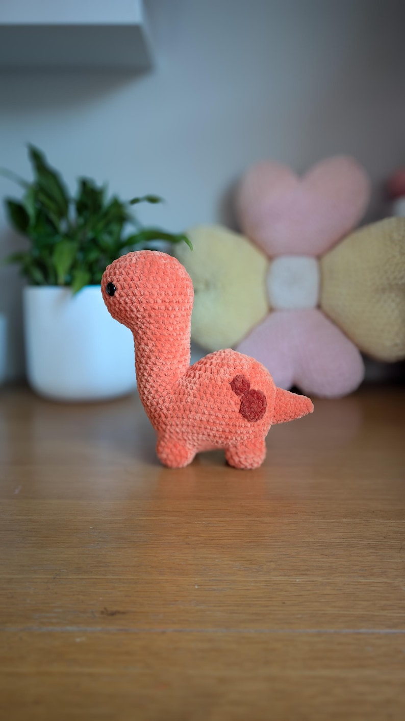 Pu&ograve; includere: Un giocattolo di dinosauro di crochet arancione con occhi neri. Il dinosauro &egrave; in piedi su una superficie di legno.