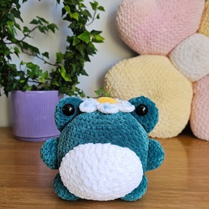 May include: A teal crochet frog plushie with a white belly and a daisy crown. The frog has large black eyes and is sitting on a wooden surface. A purple pot with greenery is in the background.