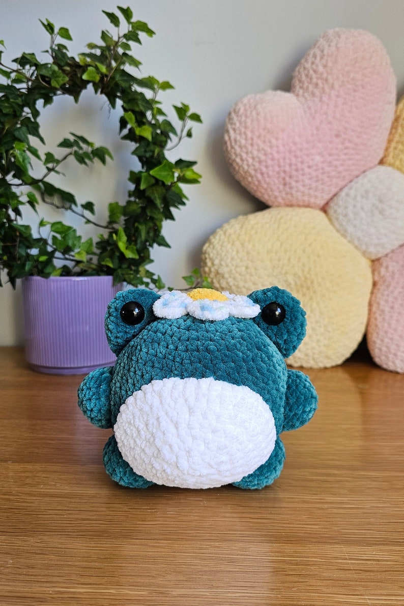 May include: A teal crochet frog plushie with a white belly and a daisy crown. The frog has large black eyes and is sitting on a wooden surface. A purple pot with greenery is in the background.