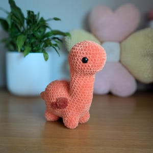 Pu&ograve; includere: Un giocattolo di dinosauro di crochet arancione con occhi neri. Il dinosauro &egrave; seduto su una superficie di legno.