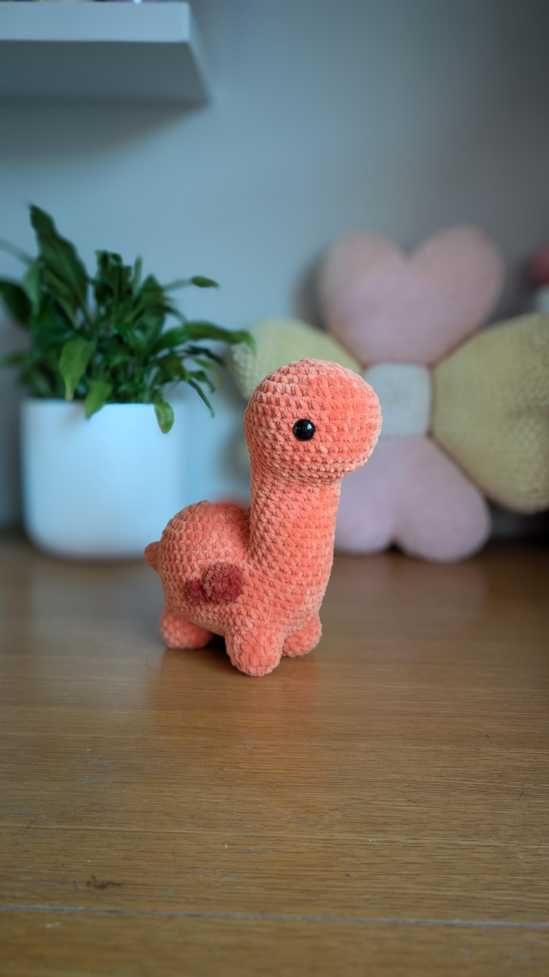 Pu&ograve; includere: Un giocattolo di dinosauro di crochet arancione con occhi neri. Il dinosauro &egrave; seduto su una superficie di legno.