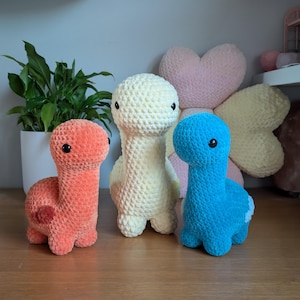 Pu&ograve; includere: Tre peluche di dinosauro all'uncinetto in diversi colori: arancione, giallo e blu. I peluche sono seduti su una superficie di legno.