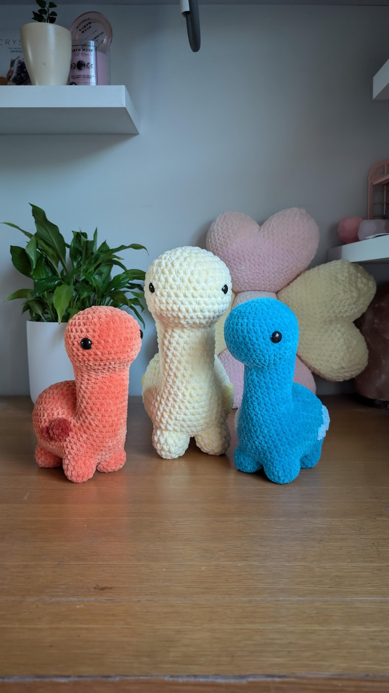 Pu&ograve; includere: Tre peluche di dinosauro all'uncinetto in diversi colori: arancione, giallo e blu. I peluche sono seduti su una superficie di legno.