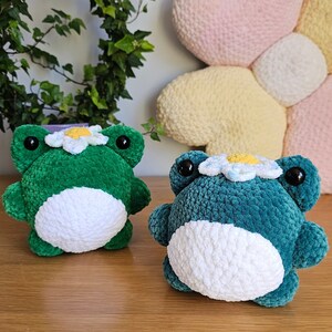May include: Two plush frog toys in the image. One is green, the other is teal. Both have white bellies, black eyes, and daisy crowns. The background includes a heart-shaped pillow and a leafy plant.
