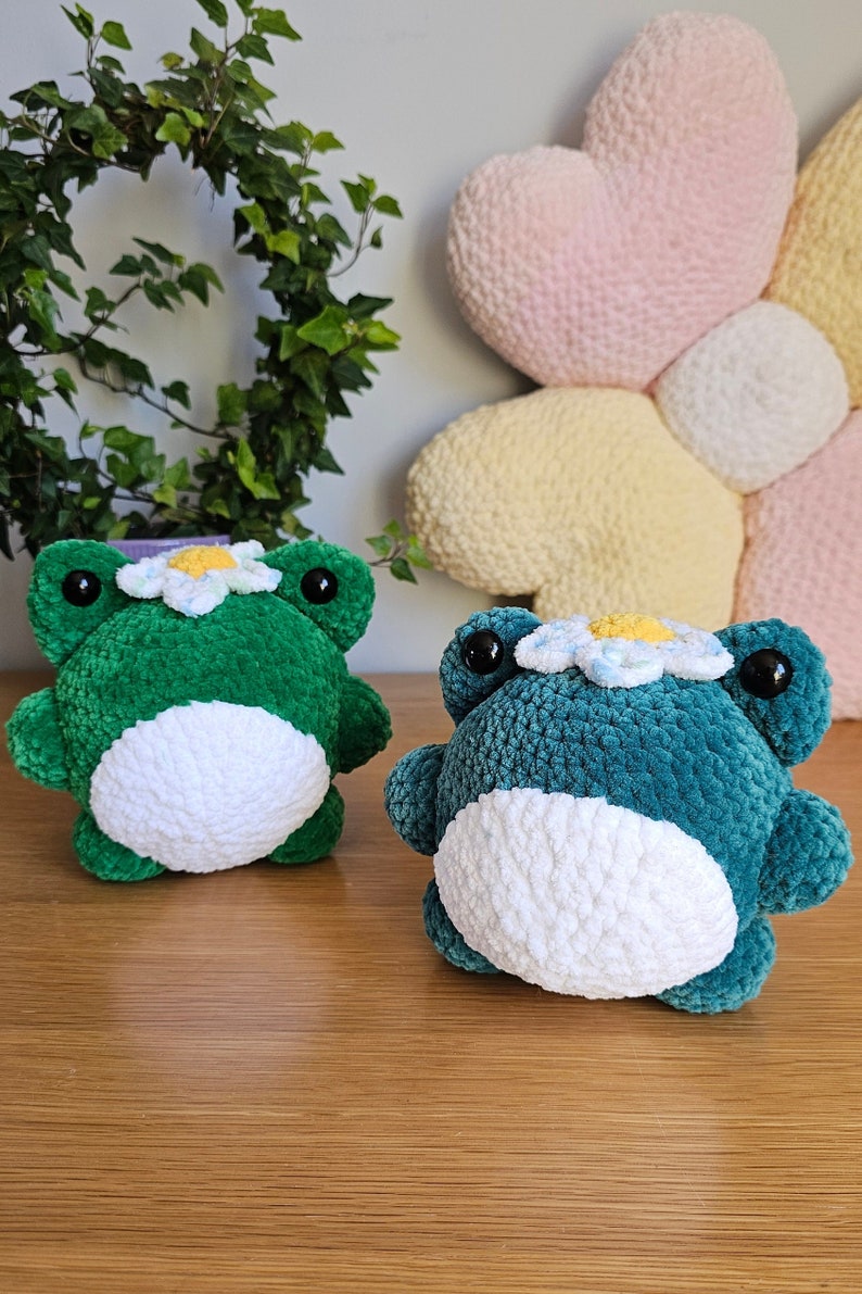 May include: Two plush frog toys in the image. One is green, the other is teal. Both have white bellies, black eyes, and daisy crowns. The background includes a heart-shaped pillow and a leafy plant.